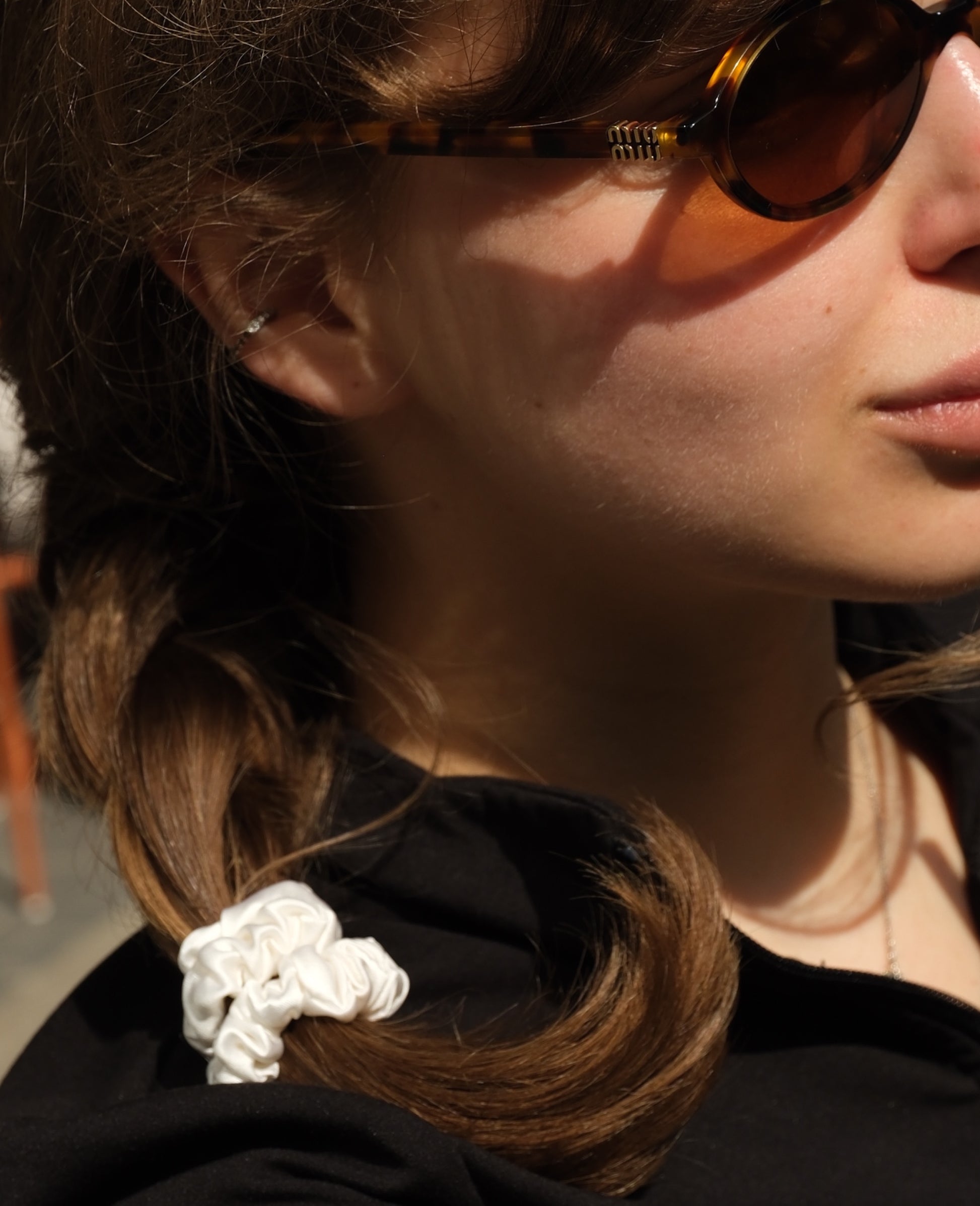 Close-up of a model wearing a white LISS silk scrunchie in sunlight, paired with tortoiseshell sunglasses